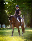 A woman riding a chestnut horse in a sunny field with a white saddle pad under the saddle.
