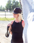 Jackie in a black Jackie&Co cap with the orange line art horse logo standing with her grey horse on a sunny afternoon in the paddocks.