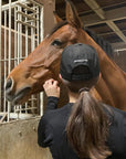 A woman with dark hair in a black Jackie&Co cap giving a brown horse affection.