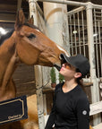 A woman with dark hair in a black Jackie&Co cap with a chestnut horse giving her a kiss in the barn.