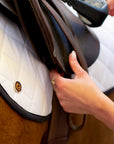Close-up view of a woman adjusting a saddle over a white Jackie&Co saddle pad on a chestnut-coloured horse.