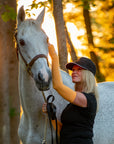 Jackie affectionately petting her grey horse's mane in orange UV protection sleeves and a black cap.