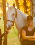 Two women in front of a grey horse wearing black Jackie&Co caps and UV protection arm sleeves in black and orange on a sunny warm day.