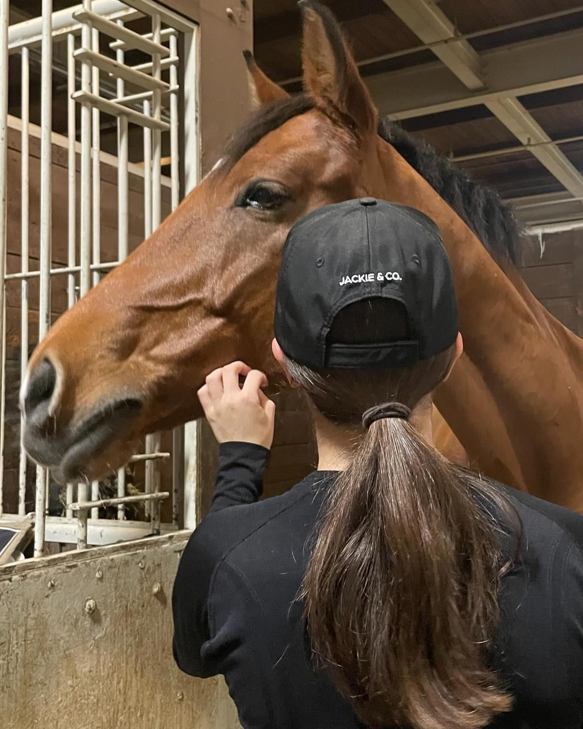 A woman with dark hair in a black Jackie&Co cap giving a brown horse affection. 