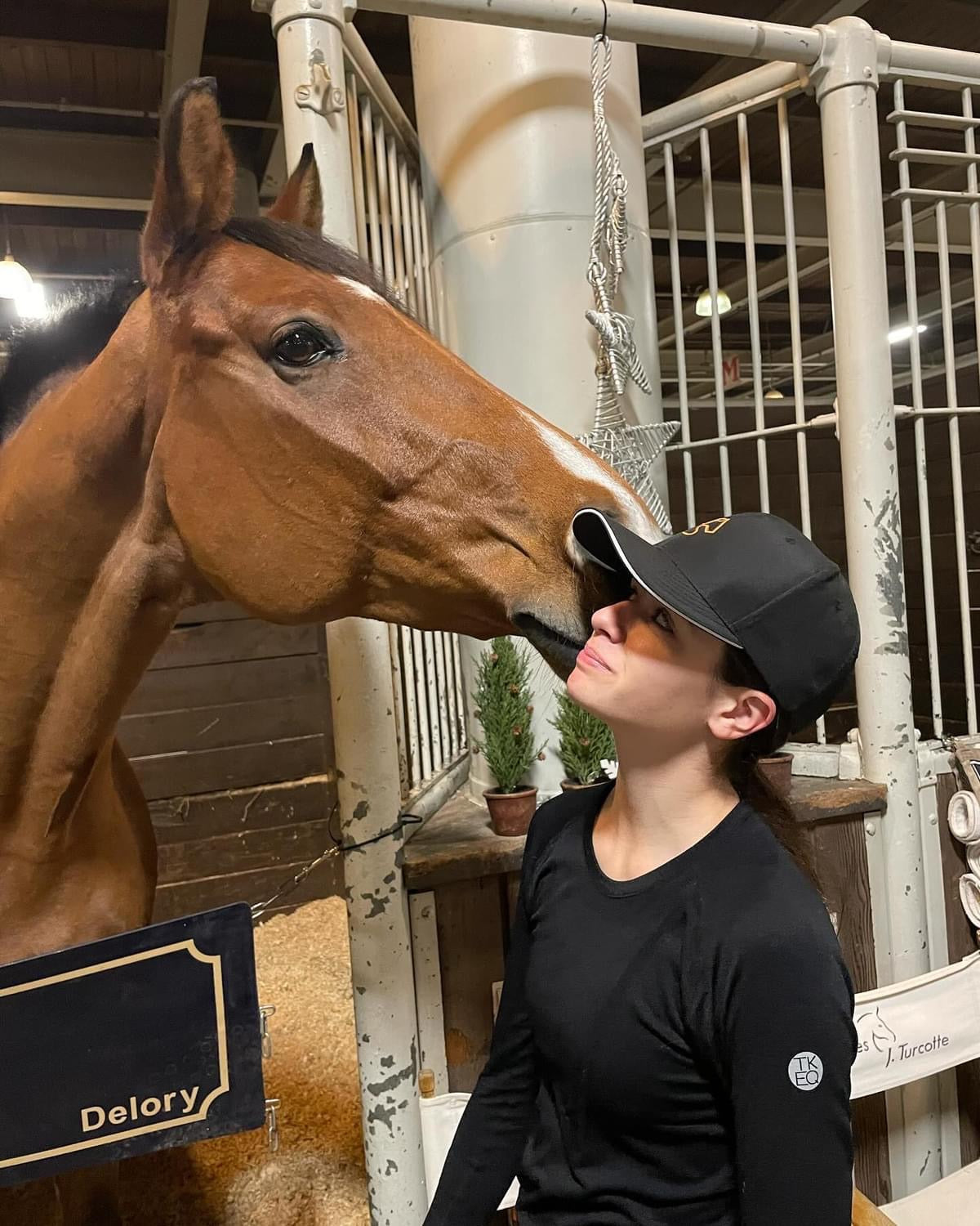 A woman with dark hair in a black Jackie&Co cap with a chestnut horse giving her a kiss in the barn. 
