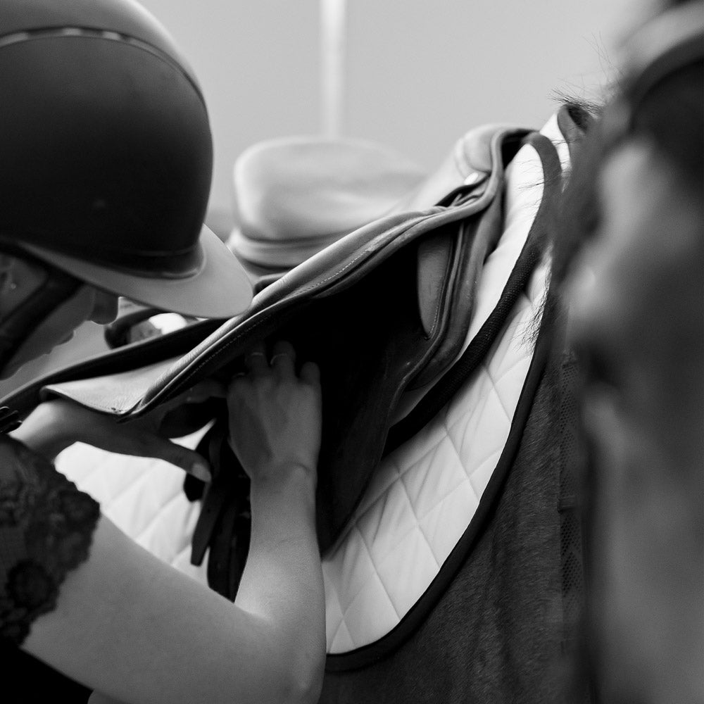 A rider adjusting the saddle with a white Jackie&Co saddle pad under it. 