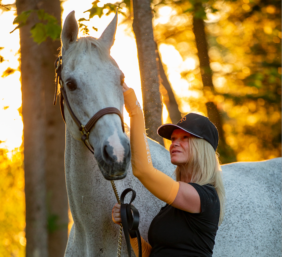 Jackie rubbing her grey horse's head affectionately while wearing a black cap and orange sleeve guards, standing in sunlight surrounded by forest trees.