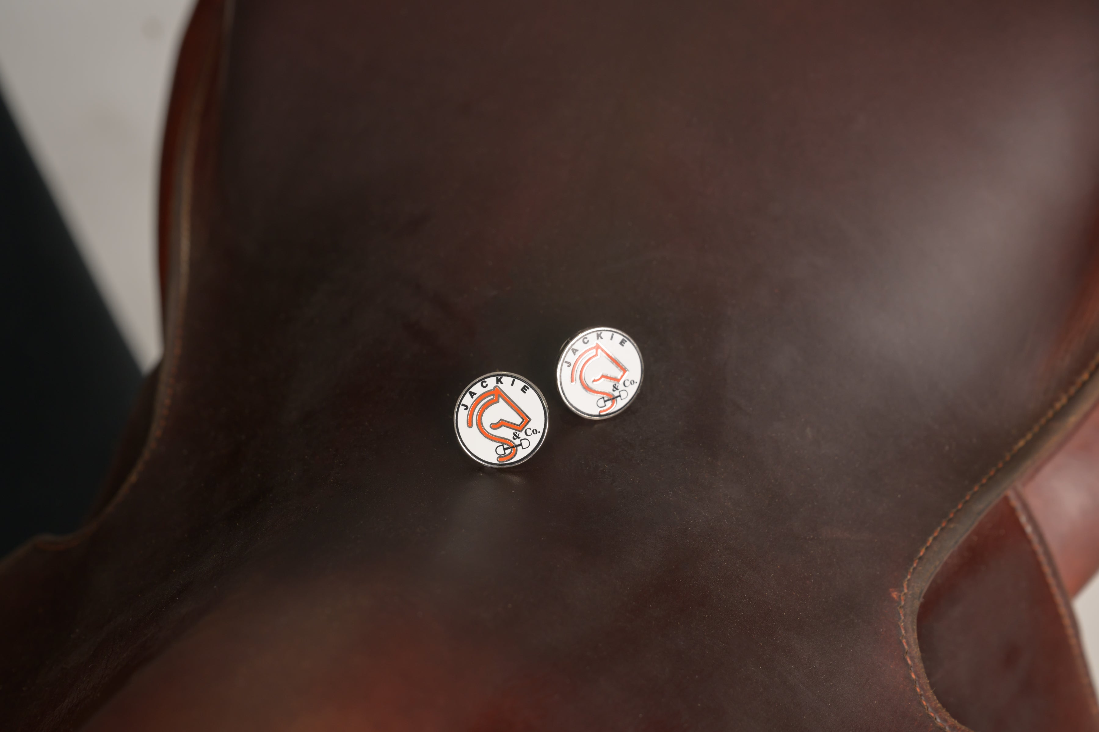 Two white Jackie&Co lucky number pins resting on a dark brown saddle. 