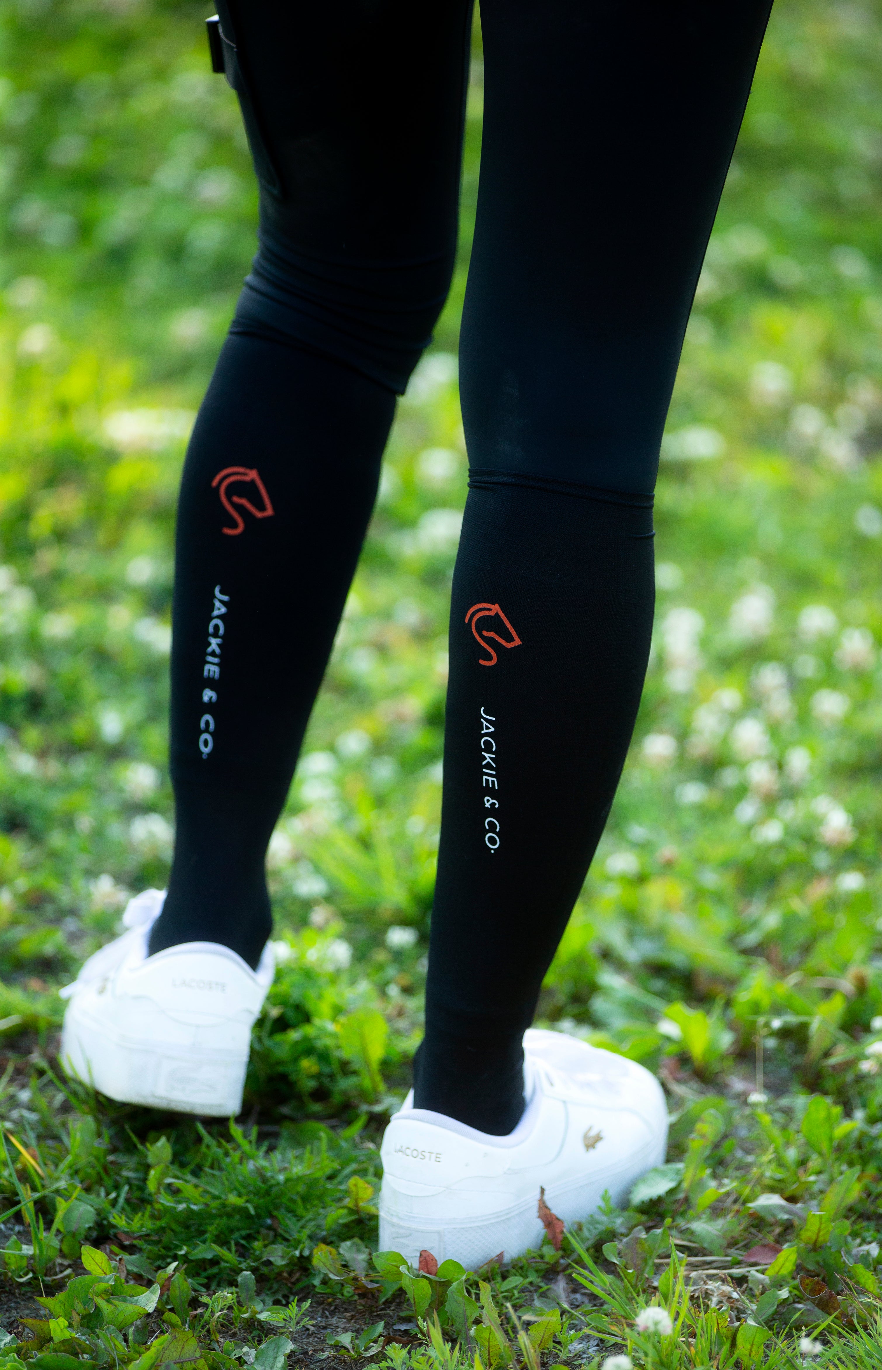 Black Jackie&Co riding socks with and orange line art horse logo on the top of the calf. Worn over black leggings with white sneakers. 